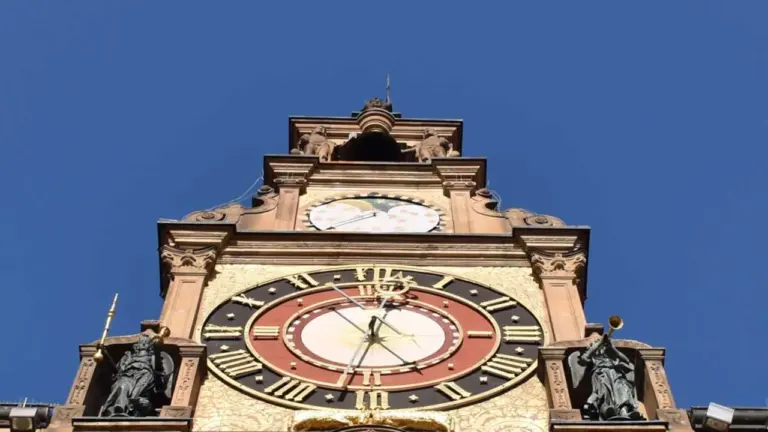 Ein historischer Uhrturm mit aufwendigen Verzierungen und zwei Engelfiguren vor einem tiefblauen Himmel.