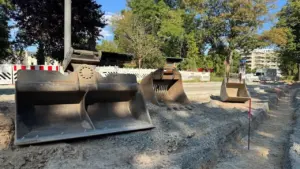 Drei verschiedene Baggerschaufeln liegen auf einer sandigen Baustelle unter blauem Himmel bereit.
