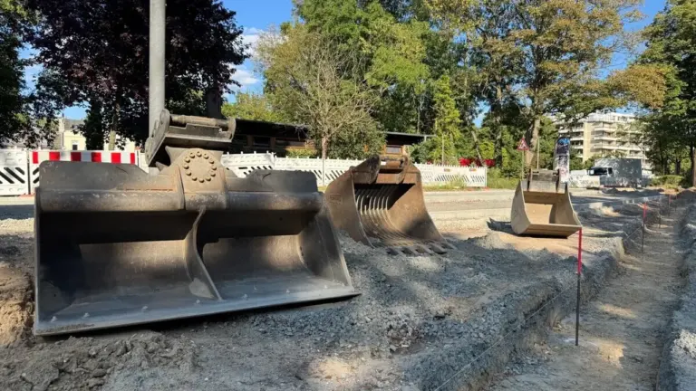 Drei verschiedene Baggerschaufeln liegen auf einer sandigen Baustelle unter blauem Himmel bereit.