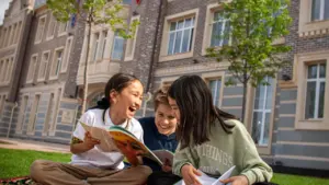 Drei glückliche Kinder lesen gemeinsam auf einer Picknickdecke vor einer Schule.