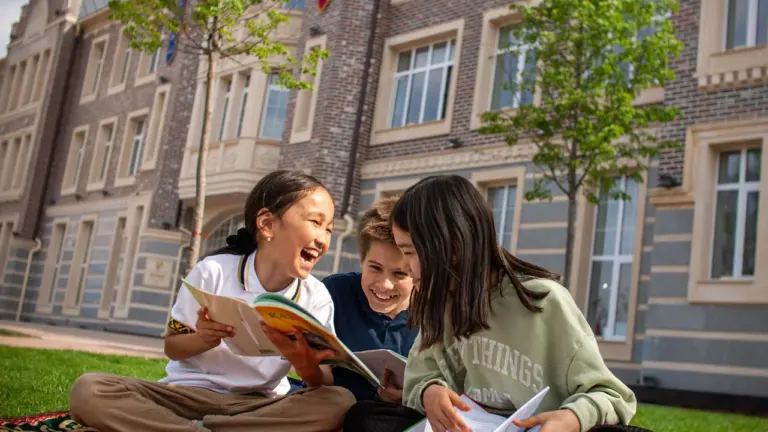 Drei glückliche Kinder lesen gemeinsam auf einer Picknickdecke vor einer Schule.