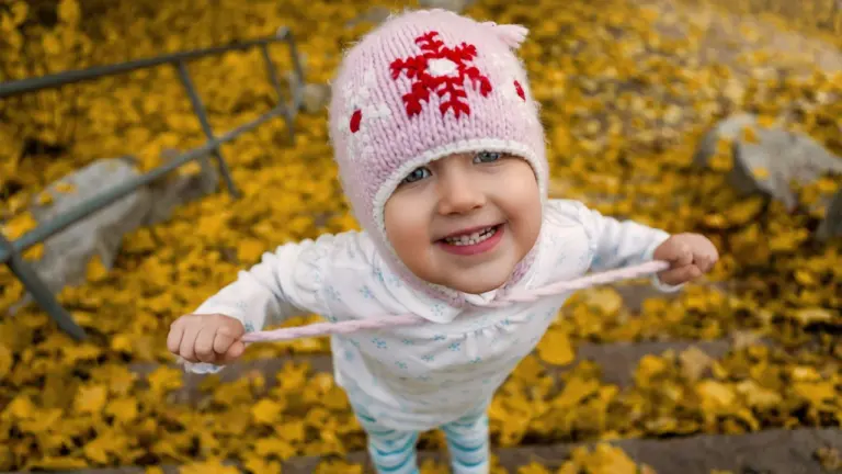 Ein kleines Kind mit rosa Mütze steht fröhlich inmitten von buntem Herbstlaub im Park.